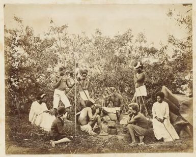 Kanak men and women relaxing around a campfire smoking pipes and holding spears, New Caledonia, ca. 1870s / Allan Hughan