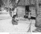 Native women in front of their home weaving palm branches, Rongerik Island, 1947