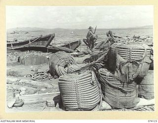 HANSA BAY, NEW GUINEA. 1944-06-17. A MEMBER OF THE 4TH INFANTRY BRIGADE SURVEYS THE SCENE OF DEVASTATION ON THE BEACH AFTER THE HEAVY ALLIED BOMBING OF THE AREA. NOTE THE LARGE BALES OF MANILLA ..