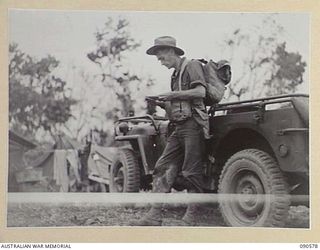 WONGINARA MISSION, NEW GUINEA. 1945-04-08. LIEUTENANT R.J. BUCHANAN, PHOTOGRAPHER, 6 MILITARY HISTORY FIELD TEAM ATTACHED HEADQUARTERS 6 DIVISION, EXAMINING PAPERS BESIDE A JEEP DURING OPERATIONS ..