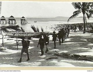 PAPUA, NEW GUINEA. 1942-07. AUSTRALIAN SOLDIERS IN A TROPICAL SETTING. AN INFANTRY PATROL PASSES ALONG THE FORESHORE BY A NATIVE VILLAGE, SEVERAL HUTS OF WHICH CAN BE SEEN STILTED OUT OF THE ..