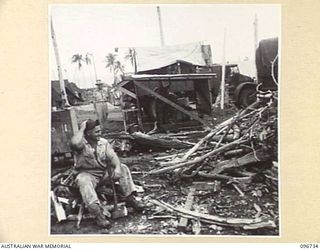 MOEM, WEWAK AREA, NEW GUINEA, 1945-09-17. CRAFTSMAN C.W. BRINGOLF, 2/4 ARMOURED REGIMENT DETACHMENT WITH 4 INFANTRY TROOPS WORKSHOP, SITTING ON CHOPPED WOOD LOOKING AT THE REMAINING PILE