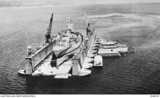 PACIFIC OCEAN. C.1945. AN AERIAL VIEW OF AN ADVANCED BASE SECTIONAL DOCK OF THE US NAVY AT A PACIFIC ISLAND BASE. THE DOCK IS SERVICING A COLORADO CLASS BATTLESHIP. NOTE THE BARGES ALONGSIDE TO ..