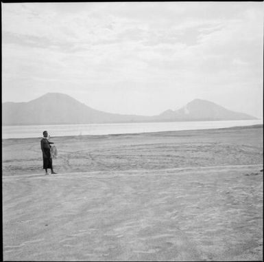 Man smoking a pipe and holding some fish on a beach with Mother and South Daughter Mountains in the distance, Rabaul Harbour, New Guinea, 1937 / Sarah Chinnery