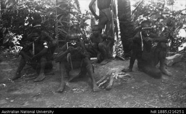 Six men sitting on the ground near trees, one man standing