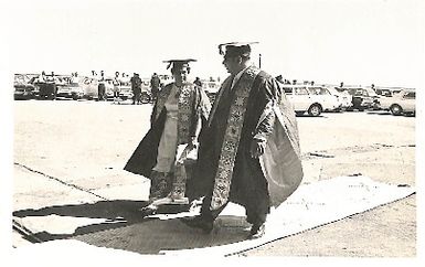 Photo of male and female dressed in graduation gowns; male walking over woven pandanus mat (67534)