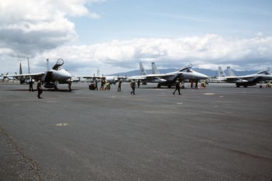 A right front view of three F-15 Eagle aircraft from the 21st Tactical Fighter Wing, parked on the flight line during Exercise OPPORTUNE JOURNEY 84. A C-130 Hercules aircraft can be seen in the background