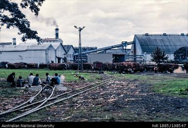 Fiji - group of workers sitting near tracks behind suger refinery, trucks holding sugarcane