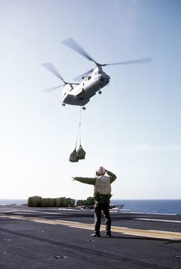 A crew member signals to the pilot of a UH-46D Sea Knight helicopter as supplies are delivered to the amphibious assault ship USS GUAM (LPH-9)