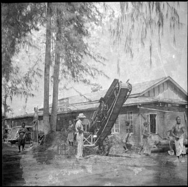 European man operating a conveyor belt and several other men with shovels clearing debris outside a store, Rabaul, New Guinea, 1937 / Sarah Chinnery
