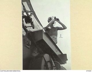 AT SEA. 1944-06-24. LIEUTENANT R.J.B. TRIMBLE, ROYAL AUSTRALIAN NAVY, USING HIS BINOCULARS TO STUDY THE SURROUNDING WATERS WHILE ON THE BRIDGE OF THE FAIRMILE ML (MOTOR LAUNCH) AS THEY PATROL ALONG ..