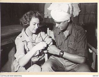 AITAPE, NEW GUINEA. 1945-03-29. MISS B. BROWN (1), A RED CROSS VISITOR TO 104 CASUALTY CLEARING STATION, SHOWS PRIVATE KARNAIL SINGH (2), HOW TO MAKE A FELT SLIPPER. HE IS RECUPERATING FOLLOWING ..