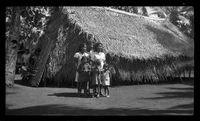 [Adults and children outside thatch building]