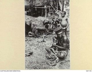 DUMPU AIRSTRIP, NEW GUINEA. 1943-11-14. A POLICE BOY NURSING A NATIVE BABY WHILE THE MOTHER WORKS IN THE 7TH AUSTRALIAN DIVISION HEADQUARTERS AREA