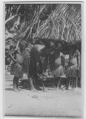 "Bock and Hessle Gilbertöarnaexpedition: 3 children and 1 young woman dressed petal skirts. The woman in the process of opening in coconut. Behind them 1 open hut. Text on the back: “The coconut is opened by pressing against an impaired pointed pouch on the ground.”: Included in series with a photon. 6977: 1-15. "