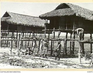 PORT MORESBY, PAPUA. 1942-07-20. AUSTRALIAN SOLDIERS LAYING BARBEDWIRE ENTANGLEMENTS THROUGH THE "STILTS" OF A NATIVE VILLAGE. COASTAL NATIVES OF NEW GUINEA BUILD VILLAGES THIS WAY BECAUSE OF AN ..