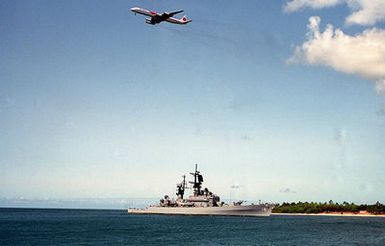 The guided missile cruiser USS WORDEN (CG-18) approaches the entrance to the Pearl Harbor channel as an airliner departing Honolulu International Airport passes overhead.