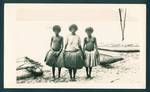 Three young New Guinean women at beach, traditional outrigger canoes in background, New Guinea, c1929 to 1932