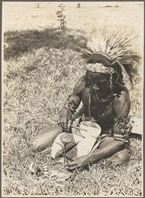 Buna Bay District [man seated on grass making a hole in a shell for a necklace] Frank Hurley