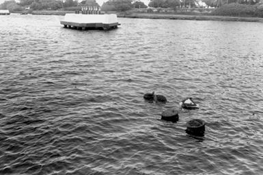 Flowers and a wreath float past the mooring bits of the sunken battleship USS ARIZONA (BB-39). In the background, a Marine honor guard waits on a mooring block to fire a salute during the ceremony commemorating the 40th anniversary of the Japanese attack on Pearl Harbor