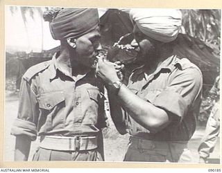 AITAPE, NEW GUINEA. 1945-03-29. SERGEANT KHAZON SINGH LIGHTING A CIGARETTE FOR SERGEANT HURDITA RAN. BOTH ARE MEMBERS OF THE INDIAN ARMY, CAPTURED WHEN SINGAPORE FELL AND FORCED TO WORK IN LABOUR ..