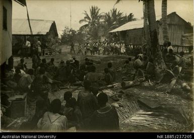 Crowd seated in foreground watching dancers and singers in space between two buildings