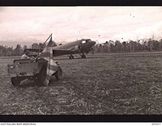 NADZAB AIRSTRIP, NEW GUINEA. 1943-09-18. THE STATION COMMANDER IN HIS JEEP, USING A BRIGHTLY COLOURED JAPANESE REGIMENTAL FLAG, TO GUIDE THE AIRCRAFT TO THEIR PARKING PLACES OFF THE STRIP