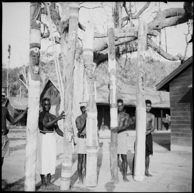 Five men each holding a carved totem pole with a building visible to the right, New Guinea, ca. 1936 / Sarah Chinnery