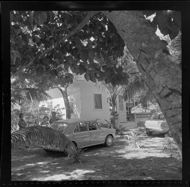 A scene in Korolevu, Fiji, showing cars including an Austin 1100 parked outside a wooden building, with two unidentified women standing alongside