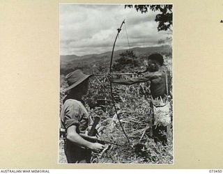 ALEXISHAFEN, NEW GUINEA, 1944-05-18. NX147907 PRIVATE F. CINCOTTA (LEFT), NO. 9 PLATOON A COMPANY, 35TH INFANTRY BATTALION, AN OWEN GUNNER, WATCHES A KANAKA NATIVE AIMING HIS BOW AND ARROW IN THE ..