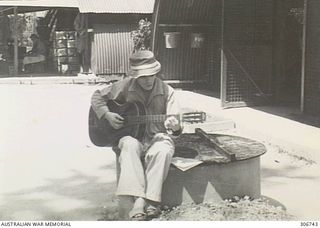 A Japanese prisoner at the RAN War Criminal Compound at the RAN shore base, sits in a courtyard playing a guitar. The instrument was supplied by the International Red Cross, Tokyo. Work on this RAN ..