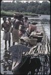 Canoes: man places basket of yams and other items onto canoe for coastal transport, women holding infants stand nearby