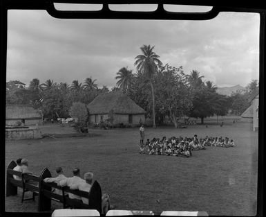 Small group of guests watching a group of children seated on the ground at a meke, Vuda village, Fiji