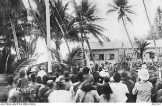 A gathering of troops, European residents and local people at the unfurling of the British flag outside the German Government Station on the surrender of Nauru.  Empty cannon shells are encircling ..