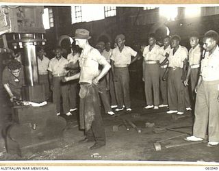 NEWCASTLE, NSW. 1944-01-28. AUSTRALIAN AND NEW GUINEA ADMINISTRATION UNIT NATIVES WATCHING EMPLOYEES OF THE BROKEN HILL PTY., WORKSHOPS FORGING A FITTING WITH A LARGE STEAM HAMMER