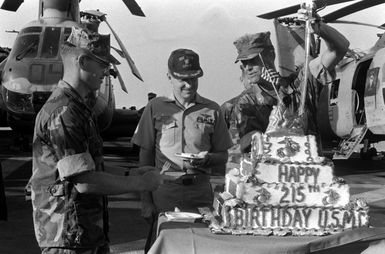 CAPT. Suffu, Commanding Officer, USS GUAM (LPH-9), is guest of honor at the Marine Corps 215th birthday ceremony near parked CH-46 helicopters on the flight deck. A Lieutenant Colonel cuts the birthday cake with a sword while another Marine looks on