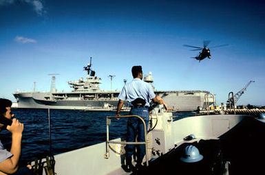 A motor whale boat from the amphibious command ship USS BLUE RIDGE (LCC-19) returns to the ship. Alongside the BLUE RIDGE is a fuel barge, and an SH-3 Sea King from the Seventh Fleet command flies overhead