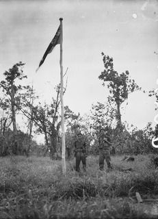 BOUGAINVILLE ISLAND. 1945-01-18. QX35765 LIEUTENANT A.E.C. MULLALY AND QX33451 PRIVATE V.M. PERROUX OF THE 47TH INFANTRY BATTALION BENEATH THE AUSTRALIAN FLAG FLYING FROM A FLAGPOLE AFTER THE ..