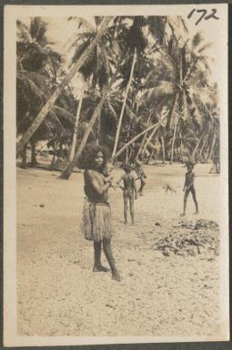 Young woman holding a baby on a beach with other children in background, Port Moresby area, Papua New Guinea, probably 1916