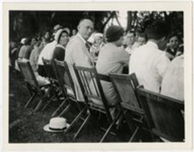Twenty or more people sitting at a long table outdoors, eating