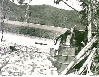 THE SOLOMON ISLANDS, 1945-10-15. A NATIVE CARRYING TWO BUCKETS ON A YOKE WITH SOME STORAGE DRUMS IN THE FOREGROUND ON A BOUGAINVILLE ISLAND BEACH. (RNZAF OFFICIAL PHOTOGRAPH.)