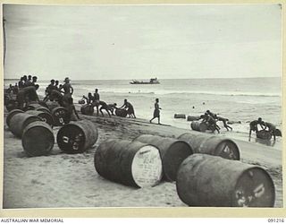 BOUGAINVILLE. 1945-04-25. NATIVES ROLLING 44-GALLON DRUMS OF FUEL ASHORE AFTER THEY HAD BEEN UNLOADED FROM BARGES INTO HEAVY SURF. A 300-TON VESSEL OF 13 SMALL SHIPS COMPANY TRANSPORTED 1200 DRUMS ..