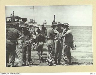 TSIMBA AREA, BOUGAINVILLE ISLAND. 1945-02-13. NX30101 MAJOR W.H. CORDELL OF AN AUSTRALIAN ARMY WATER TRANSPORT UNIT, CHECKING WITH THE BEACHMASTER WHILE NATIVES LOAD HIS BARGE WITH SUPPLIES AT PUTO