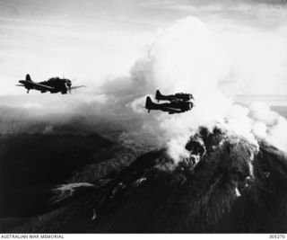 BOUGAINVILLE. C. 1944. AERIAL PHOTOGRAPH OF THREE DOUGLAS SBD DAUNTLESS DIVE BOMBER AIRCRAFT PASSING THE ACTIVE VOLCANO OF MOUNT BAGANA ON A BOMBING OPERATION AGAINST KAVIENG. (NAVAL HISTORICAL ..