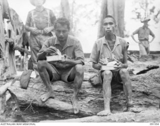 SORAKEN AREA, BOUGAINVILLE. 1945-04-03. AMBOINESE WHO SURRENDERED TO OUR TROOPS, SIT ON A LOG OUTSIDE THE 11 INFANTRY BRIGADE COOKHOUSE, HAVING THEIR FIRST SUBSTANTIAL MEAL FOR SEVERAL DAYS. THEY ..