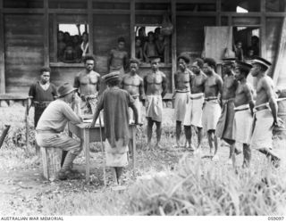 BUKAUA, NEW GUINEA. 1943-10-18. NX155085 CAPTAIN R. G. ORMSBY OF THE AUSTRALIAN AND NEW GUINEA ADMINISTRATIVE UNIT TALKING TO THE NATIVE VILLAGERS. SHOWN: VILLAGE LULUAI (CHIEF) - CAP WITH ONE ..