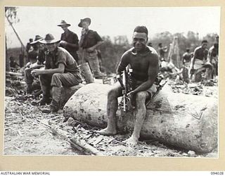 WEWAK AREA, NEW GUINEA, 1945-07-14. POLICE BOY "MARIE GOMMAH SEATED ON A GARAMUT, A NATIVE SIGNAL DRUM. HE HAS USED IT TO SIGNAL NATIVES IN THE SAURI VILLAGES THAT "THE BLOT" HAS FALLEN. IT WAS ..