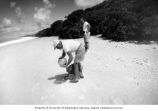 Lauren R. Donaldson picking up a glass ball from Bikini Island beach, Hill Williams stands, Summer 1964