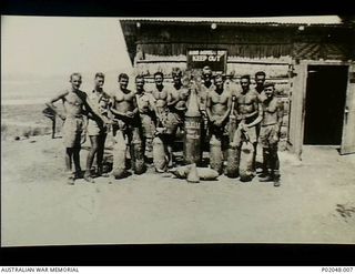 Port Moresby, New Guinea. 1943. Group portrait of all the members of a RAAF Bomb Disposal Unit standing with unexploded bombs (UXBs), in front of a shed with a sign stating "Bomb Disposal Sqn. Keep ..