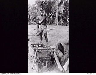 MAPRIK, NEW GUINEA. C.1946. SERGEANT N. (NAT) F. DANIELL OF THE FRONT LINE BROADCASTING UNIT, FAR EASTERN LIAISON OFFICE, SETTING UP A BATTERY POWERED FIELD AMPLIFIER UNIT, WATCHED BY A NATIVE ..